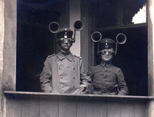 Two soldiers in historical military uniforms standing on a balcony, wearing unusual helmet-like devices with large horn-like attachments.