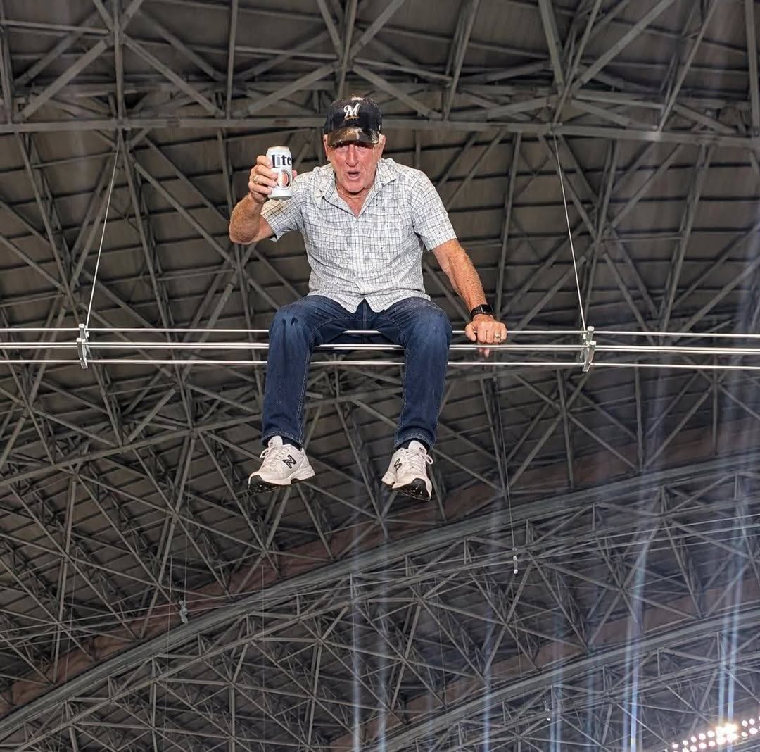 Man sitting on a beam in a sports arena, holding a beer and wearing a baseball cap, with a surprised expression against a backdrop of a large metal roof.