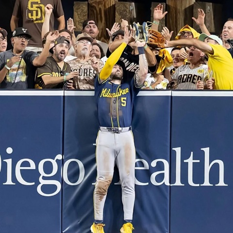 A baseball player in a blue and yellow uniform catches a fly ball while jumping against a wall, with enthusiastic fans cheering in the background.